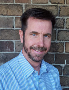 Mike is smiling. He has short brown hair, a beard and is wearing a blue collared shirt.